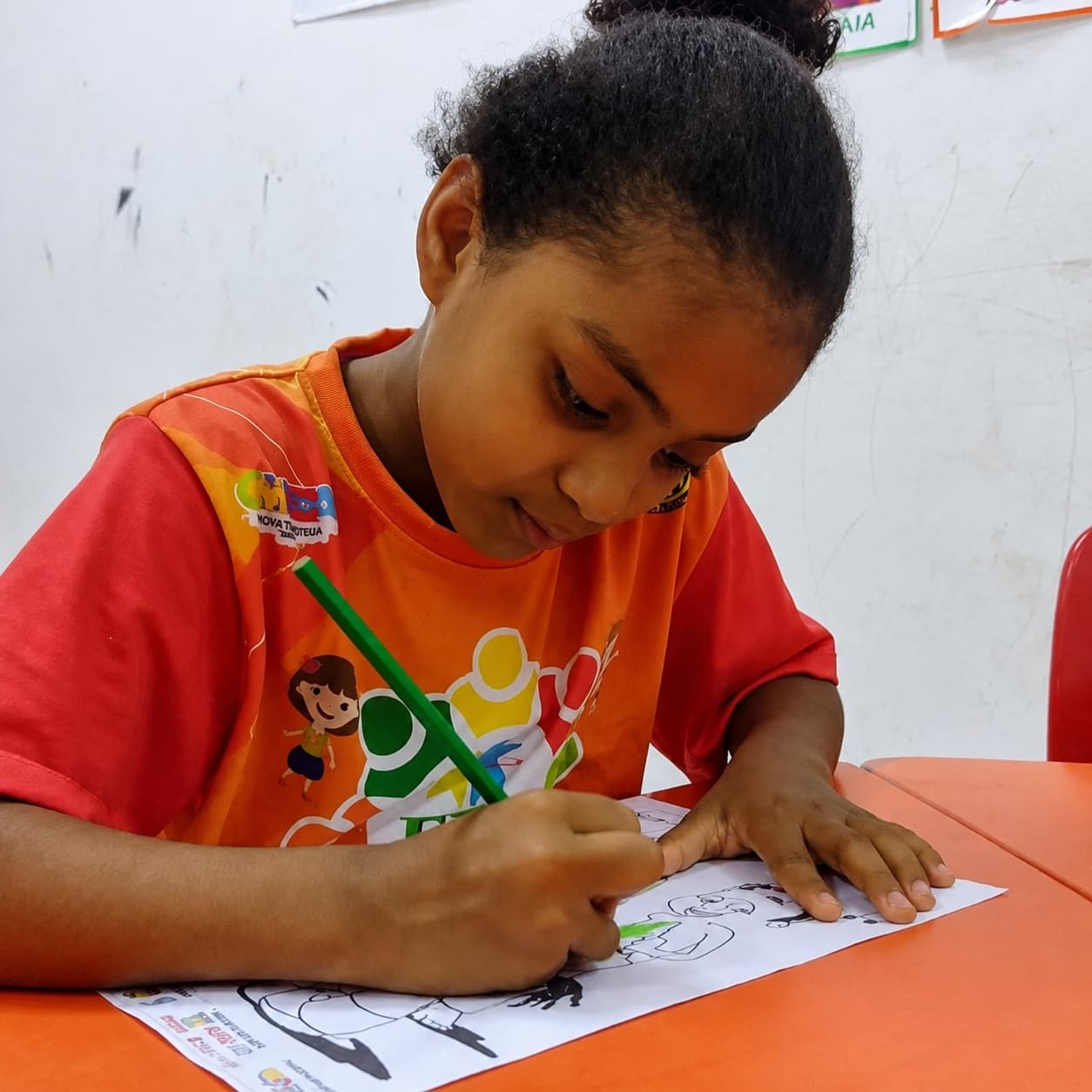 Menina concentrada escrevendo com um lápis, vestindo camiseta laranja do projeto FFC.